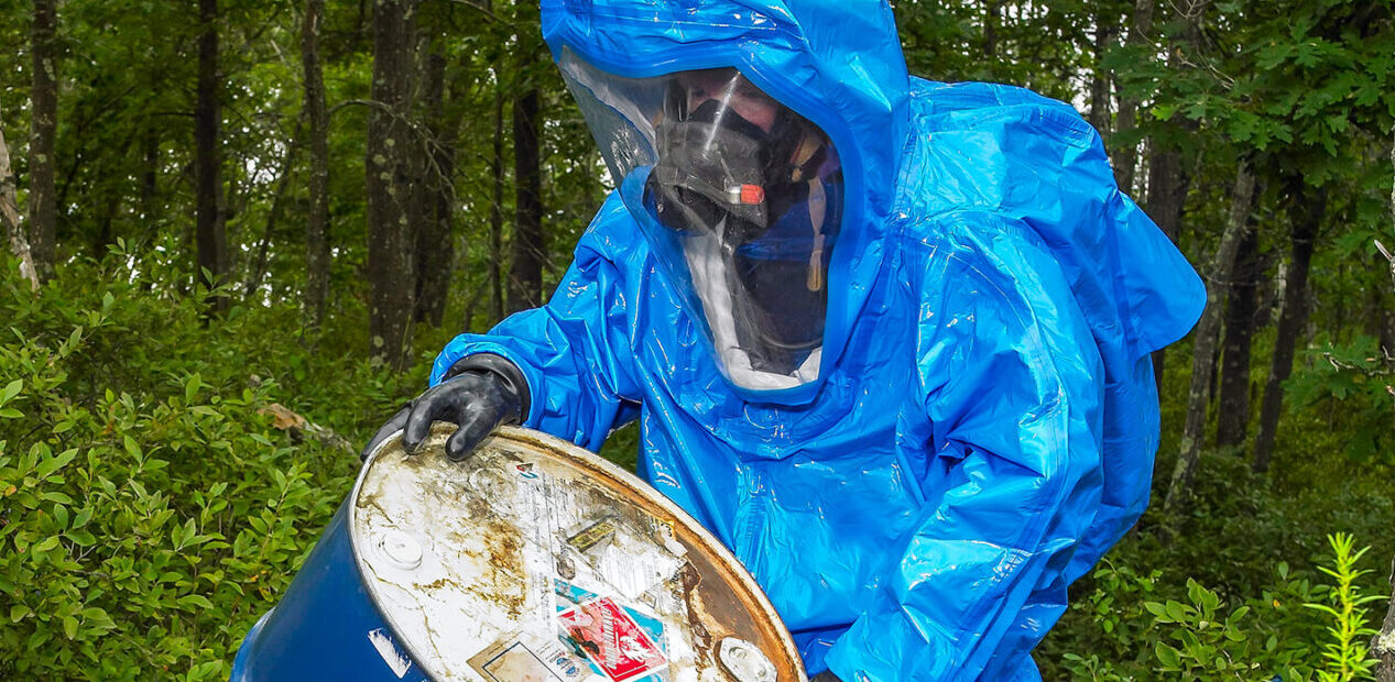 A worker wearing a Lakeland Interceptor chemical protective suit and full-face respirator handles a chemical barrel in an industrial setting. The fully encapsulating suit provides protection while the worker inspects or manages the drum as part of hazardous materials operations.